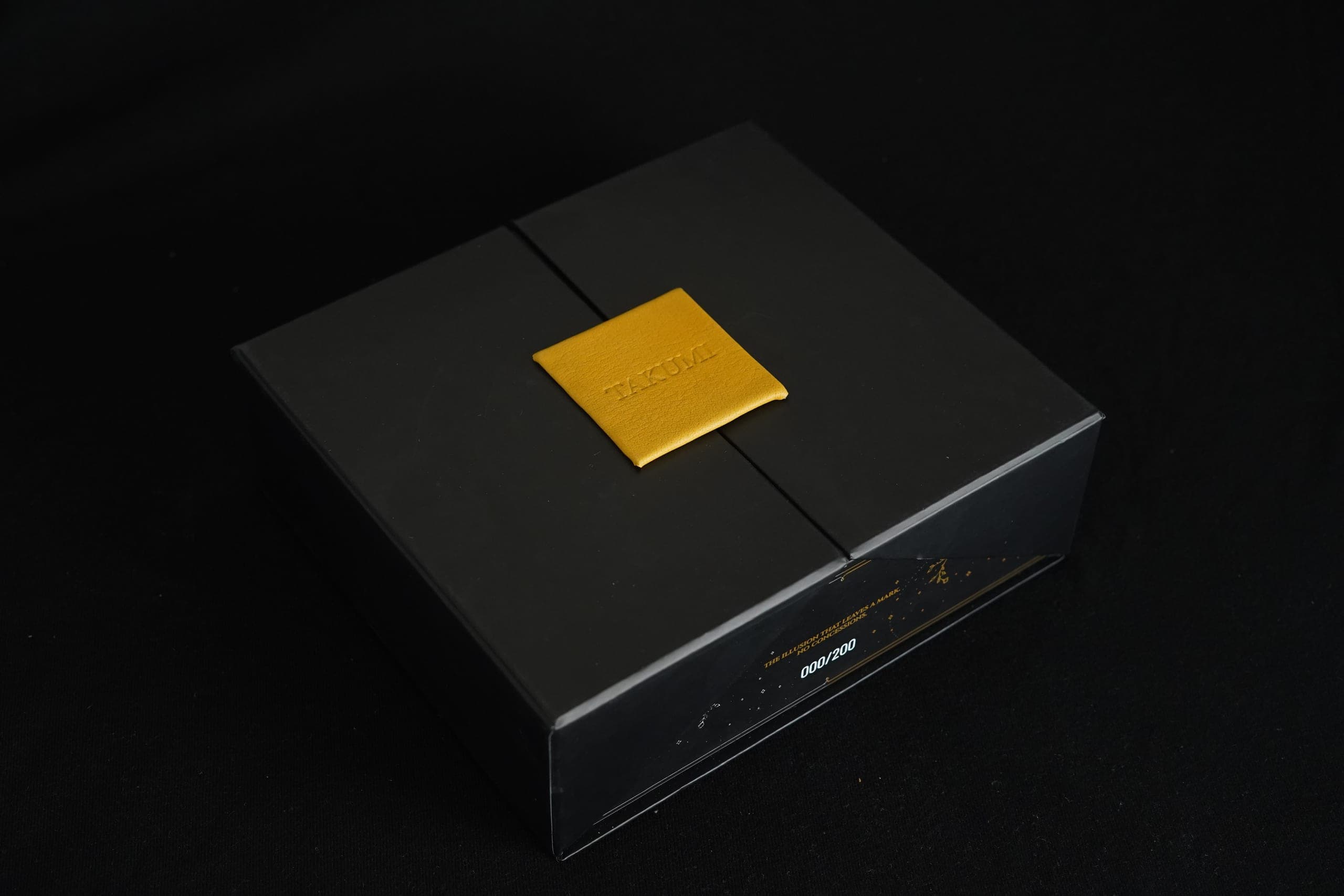 TAKUMI GENSŌ — the numbered black presentation box on deep black, gold TAKUMI leather seal centred on the lid, edition 000/200 embossed on the front edge