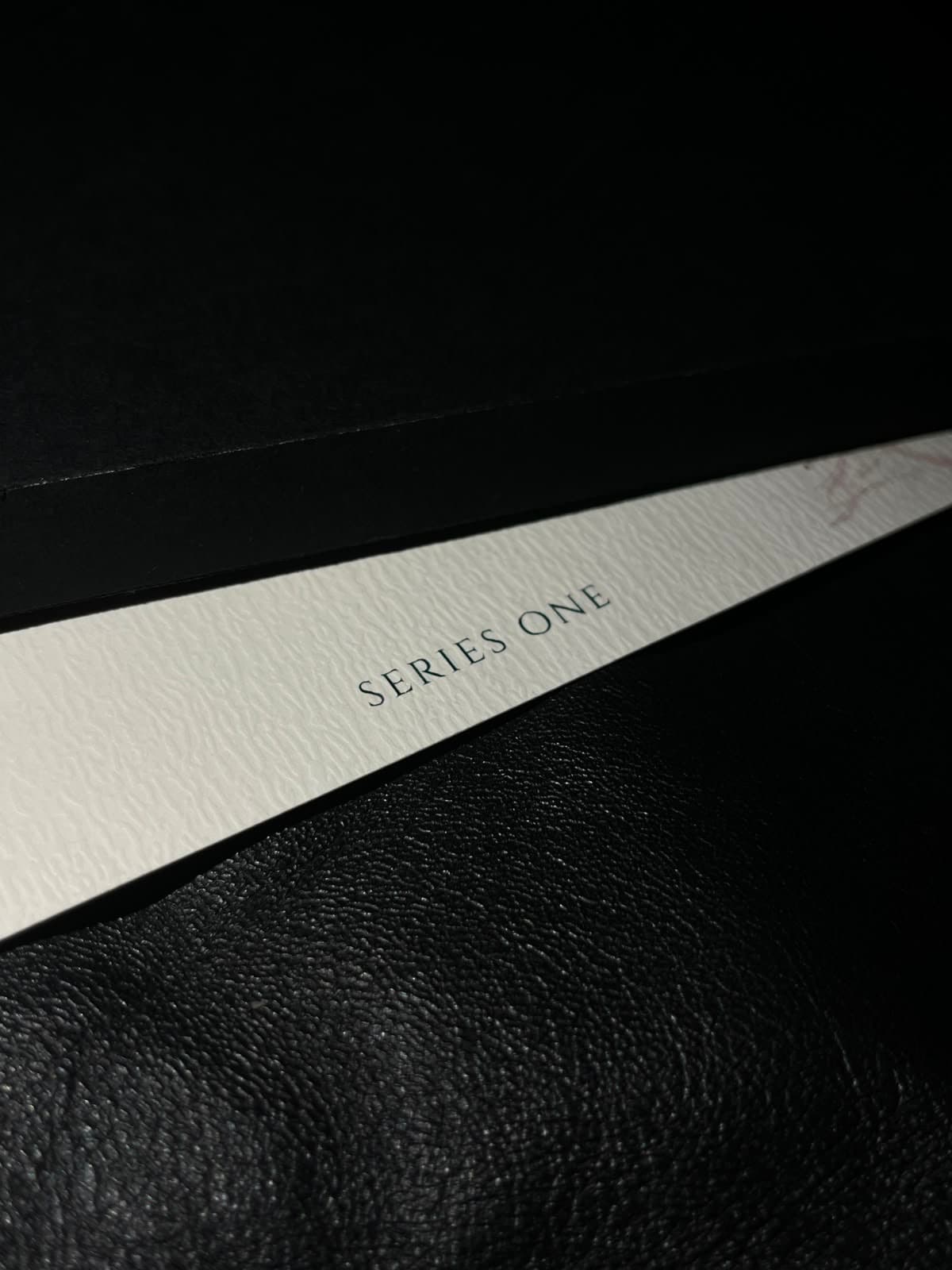 TAKUMI — Series One cards in cream textured stock on black leather, mauve line art and gold wordmark under a low work lamp