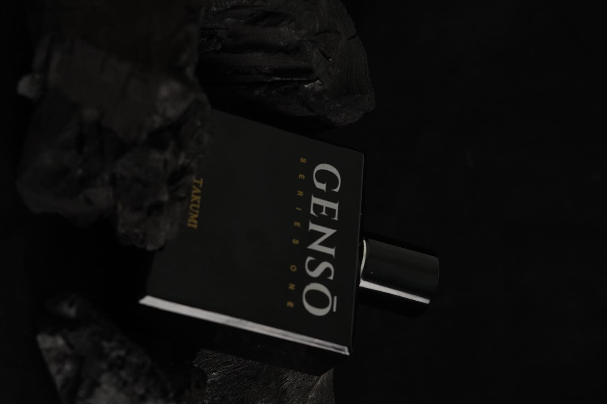 TAKUMI GENSŌ — the gloss-black bottle resting among black charcoal, the gold GENSŌ wordmark and chrome atomizer catching low light