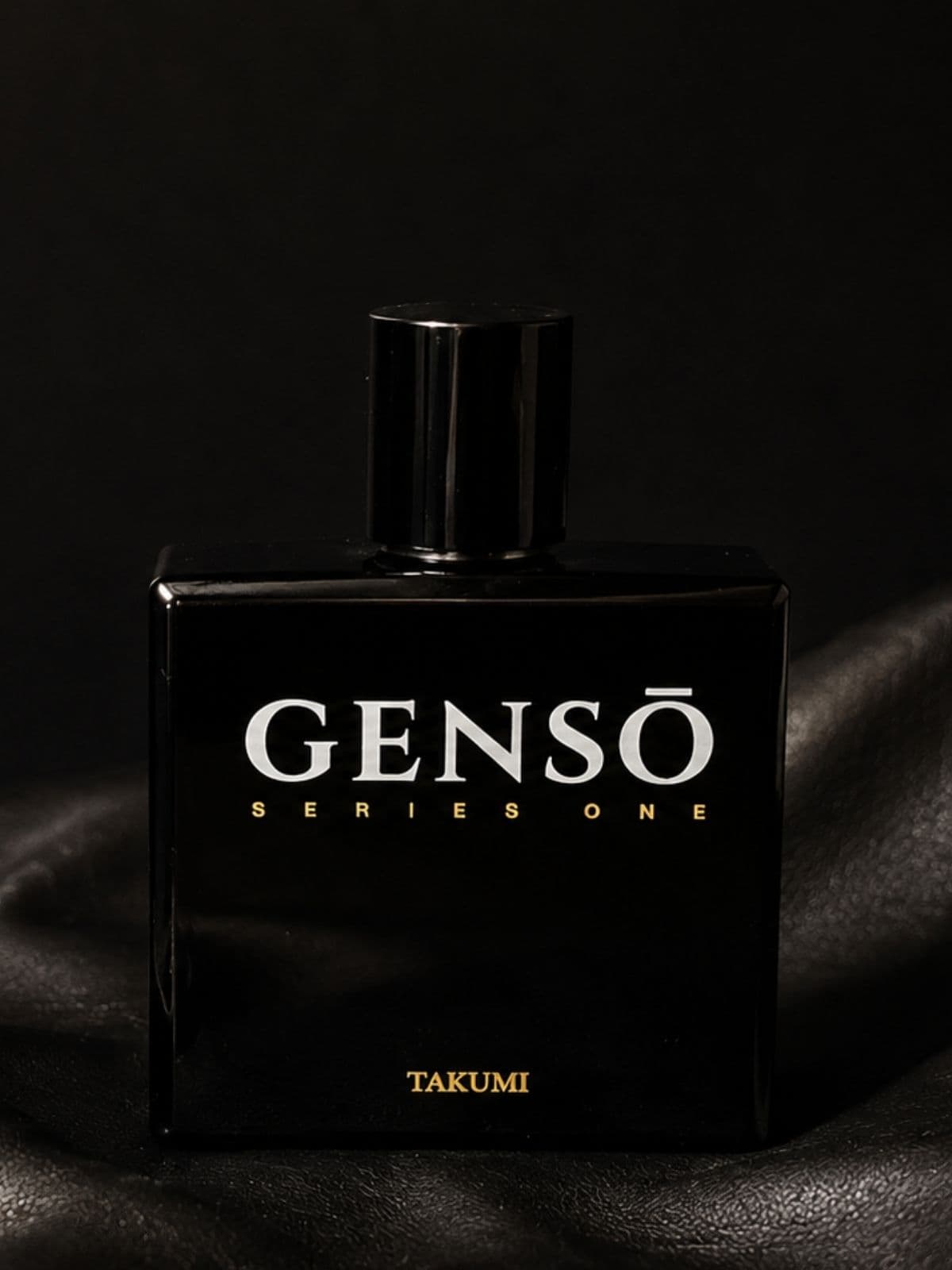 TAKUMI GENSŌ — gloss-black bottle upright in shadow, chrome atomizer exposed, cap resting beside it