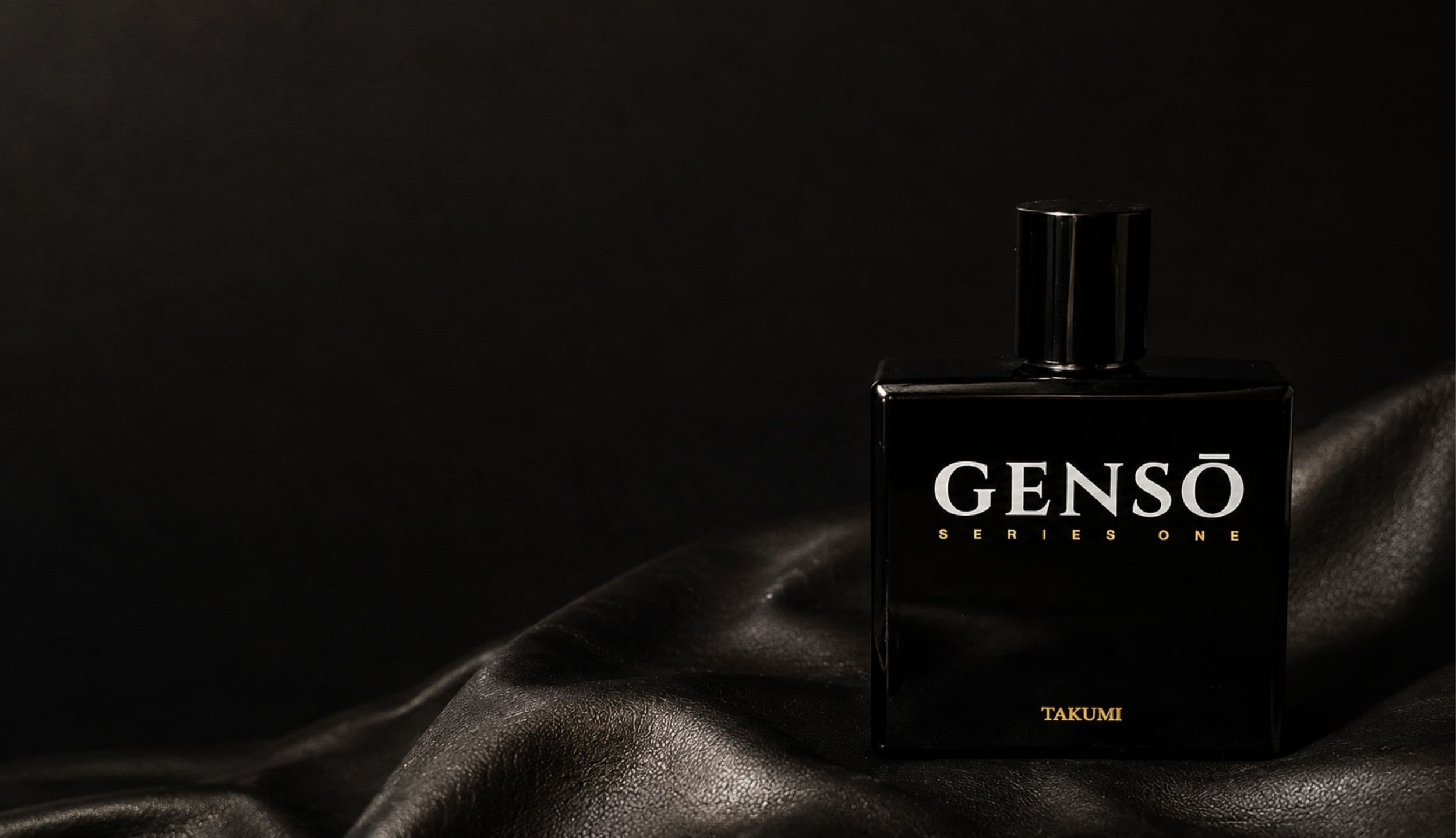 TAKUMI GENSŌ — gloss-black bottle in low light, chrome atomizer exposed, GENSŌ wordmark catching a thin edge of light