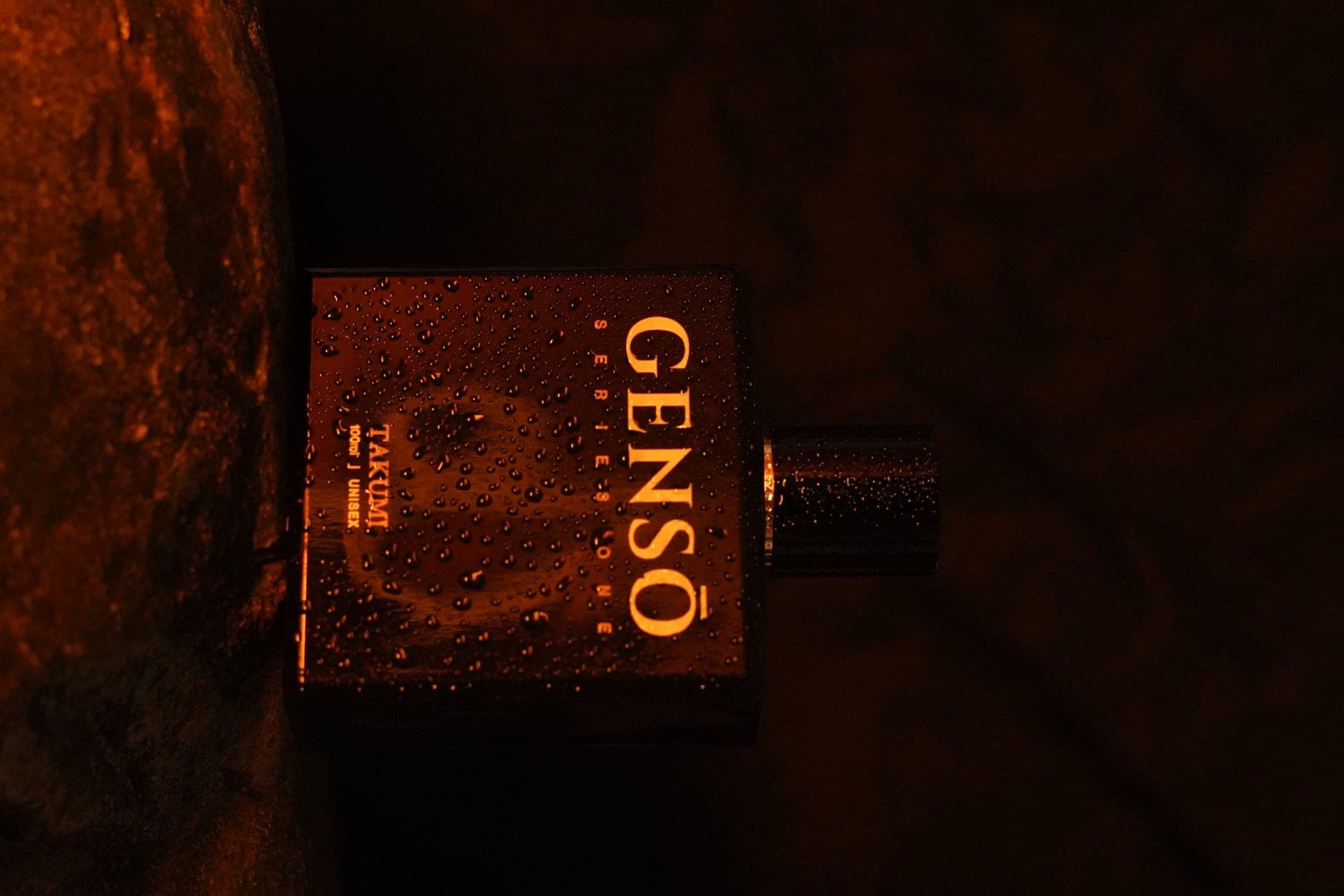 TAKUMI GENSŌ — the bottle beaded with condensation against warm amber-lit cave stone, rain before it falls